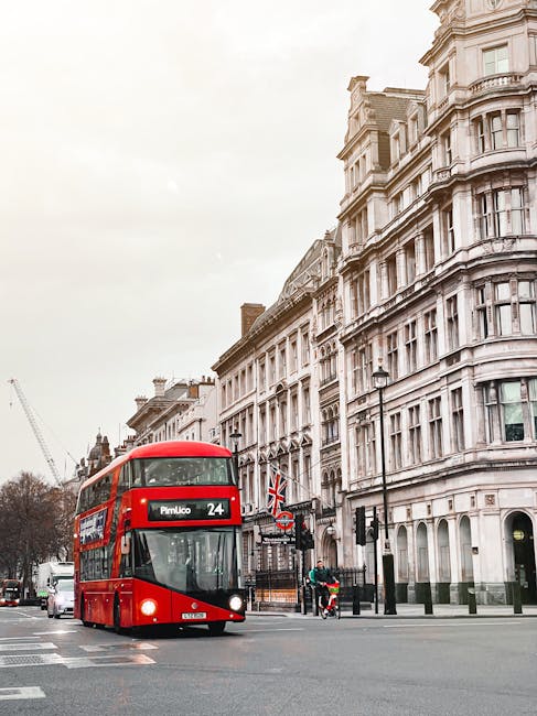 A red double-decker bus displaying the route number 24 and destination 'Pimlico' is traveling along a city street lined with tall, ornate white stone buildings that feature large windows and decorative architectural details. The bus is approaching a pedestrian crossing where a person on a bicycle and another individual are visible nearby, waiting or crossing the road. In the background, construction cranes are visible, indicating ongoing development. The scene is set during daytime with overcast weather, and the street includes black lampposts, traffic lights, and a Union Jack flag hanging from a building. The image captures a typical urban setting suitable for house removals or home relocation services, emphasizing the logistical environment of moving in the Pimlico area, with visible elements such as the bus that suggest transportation and transit in relation to furniture transport and packing and moving activities. Man and Van Pimlico occasionally operates within such environments, supporting efficient relocation and removals.