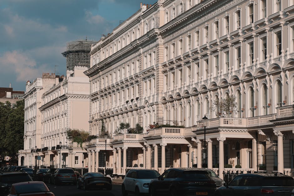 A row of white Victorian-style terraced houses with ornate architectural details, including arched windows, decorative cornices, and balconies with metal railings, situated along a street in Pimlico. The buildings are set close together, with some having small front gardens and potted plants visible on the balconies. In the foreground, a line of parked cars extends along the pavement, with a black van and a few smaller vehicles. The scene is captured during daylight with a partly cloudy sky overhead. The location is a residential area known for its elegant Georgian and Victorian properties, often included in home relocation and furniture transport services such as those provided by Man and Van Pimlico, which specialise in removals and packing during house moves.