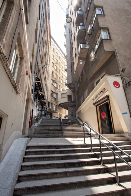 A narrow staircase with concrete steps and metal handrails ascending between high-rise residential buildings in Pimlico. The buildings have various small balconies and external air conditioning units. To the right of the staircase, there is a white, angular commercial unit with a red logo, likely part of a retail or service outlet, built partially into the building façade. The area is lit by natural daylight, with the sky visible at the top of the alley. The staircase and surrounding environment suggest a tight-access urban location suitable for small-scale home relocation and furniture transport services, as provided by Man and Van Pimlico. This scene illustrates the complex logistics involved in moving items through confined residential areas with limited access.