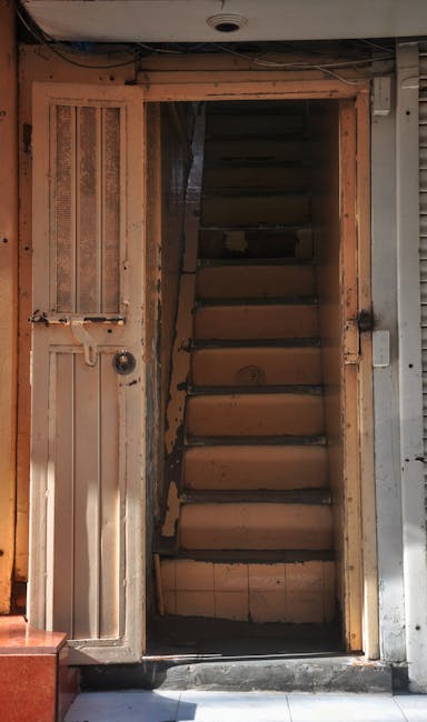A narrow wooden staircase inside a property, viewed through a doorway that has an aged and unfinished appearance with visible wood framing. The stairs are constructed of dark brown, worn wooden steps, and they ascend to an upper floor, with no handrails or banisters visible. To the left of the stairs, there is a small vintage wooden cabinet with slatted doors and a metal handle, placed against the wall. The area appears to be part of a home undergoing renovation or preparation for a house removal, illustrating the challenging access often encountered in tight-access flats. The lighting is natural, coming from outside through the doorway, which is supported by the context of a professional house removal carried out by Man and Van Pimlico, demonstrating a typical scene for furniture transport through compact stairways during home relocations.