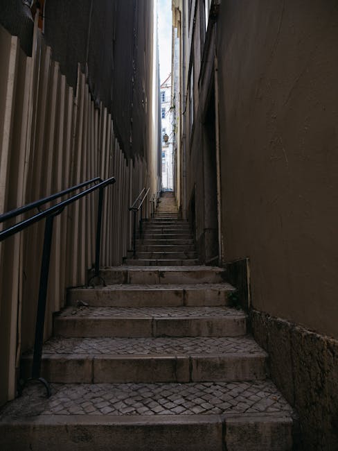 A narrow staircase with concrete steps and metal handrails ascending between high-rise residential buildings in Pimlico. The buildings have various small balconies and external air conditioning units. To the right of the staircase, there is a white, angular commercial unit with a red logo, likely part of a retail or service outlet, built partially into the building façade. The area is lit by natural daylight, with the sky visible at the top of the alley. The staircase and surrounding environment suggest a tight-access urban location suitable for small-scale home relocation and furniture transport services, as provided by Man and Van Pimlico. This scene illustrates the complex logistics involved in moving items through confined residential areas with limited access.