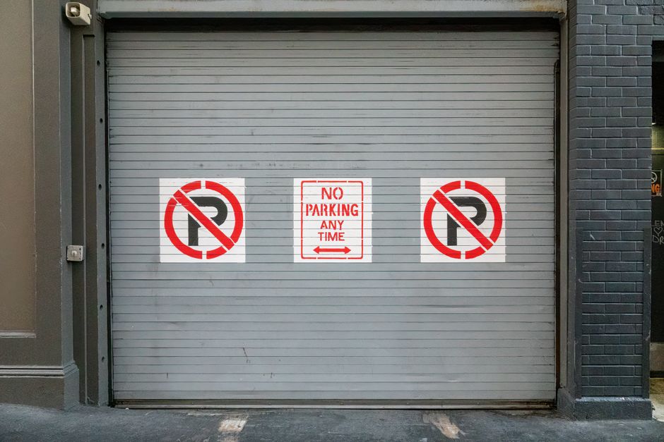 A large grey metal loading bay shutter door with horizontal slats, located outdoors and surrounded by dark brick walls. In the centre of the door, there is a red and white sign stating 'NO PARKING ANY TIME.' To the left and right of this sign, there are two additional notices featuring red circle and slash symbols over the letter 'P,' indicating no parking zones, each on a white background. The surrounding area appears to be part of a commercial or residential building, with the loading bay set at ground level. The surface in front of the door is paved, without vehicles or other objects directly in front of the shutter, suggesting a designated unloading area typical for house removals or furniture transport processes. This image is relevant for understanding loading bay restrictions and guidelines applicable to house removals or relocation services provided by Man and Van Pimlico, as outlined in the Pimlico loading bay rules and fines page.