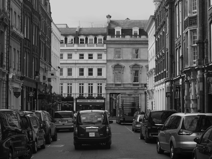 A black and white photograph of a narrow urban street in Pimlico, SW1V, showing parked cars lining both sides of the road in front of multi-storey residential buildings with classic architectural details. In the foreground, several vehicles including a small van are parked close to the pavement, with a larger delivery truck further down the street, indicating potential furniture transport or home relocation activity. The street's surface is paved, and the surroundings include windows and decorative facades typical of London townhouses. The image captures the quiet, confined setting often associated with house removals or moving logistics in a busy city area, emphasizing the need for professional removals services such as those provided by Man and Van Pimlico, to efficiently manage packing, loading, and transportation within such environment.
