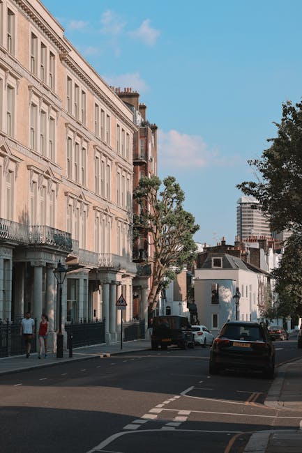 A street scene in Pimlico, London, with a row of elegant, multi-storey terraced buildings featuring large sash windows, decorative stone facades, and wrought iron balconies. The pavement is occupied by pedestrians and parked cars, while a few vehicles are seen driving along the road. Tall trees line the sidewalk, providing some greenery and shade. In the background, a modern high-rise building is visible under a blue sky with scattered clouds. This urban environment depicts a residential area suitable for house removals and furniture transport, with the setting suggesting the context of home relocation or packing and moving processes, as handled by services like Man and Van Pimlico.