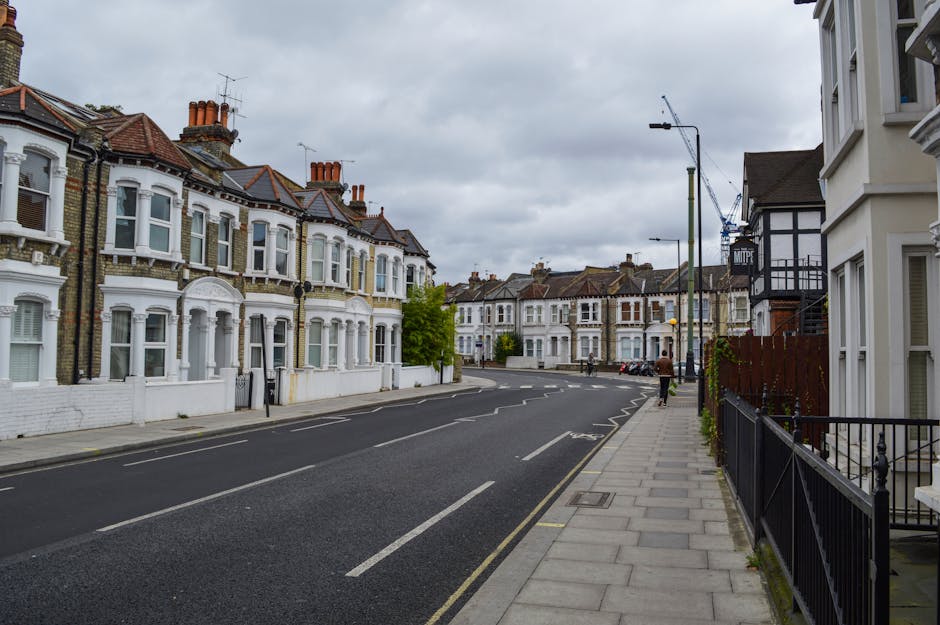 A residential street scene featuring white Victorian-style terraced houses with ornate facades, black iron balconies, and flagpoles displaying the Union Jack and a black flag. The street is lined with