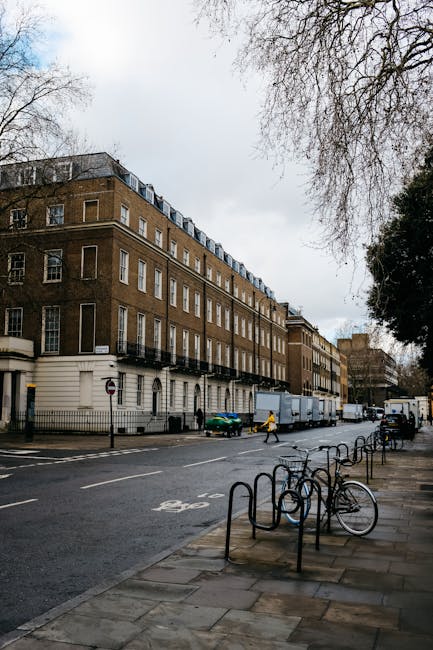 A multi-storey brick residential building with white window frames and balconies is situated along a city street in Pimlico, London. The street features several parked vehicles, including a white delivery van and a small green car, with a person walking nearby. In the foreground, a bike rack with several empty bicycle stands is positioned on the sidewalk next to a few bicycles. The sidewalk is paved with stone tiles, and a leafless tree extends its branches overhead, indicating a colder season. The overcast sky casts diffused light across the scene. This setting illustrates the typical urban environment where professional house removal services, such as those offered by Man and Van Pimlico, facilitate swift home relocations and furniture transport within the area. The image captures the process of loading or unloading furniture or packed boxes onto or from the delivery van, highlighting the logistics involved in same-day removals under time constraints, with mentions of packing, moving equipment, and on-street vehicle access.