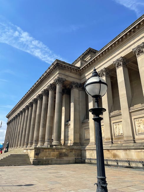 A historic neoclassical building with tall, evenly spaced Corinthian columns supporting a decorative entablature, set against a bright blue sky with a few scattered clouds. In the foreground, there is a black, vintage-style street lamp with a rounded glass lantern mounted on a ribbed pole, positioned on a paved area with stone steps leading up to the building's entrance. The scene is captured during daylight, highlighting the detailed architectural elements and the clean, orderly exterior environment. This image relates to house removals and moving services, such as those provided by Man and Van Pimlico, illustrating a typical cityscape where furniture transport and packing logistics might occur during a house relocation process from or to iconic locations like Tate Britain, near St George's Square.