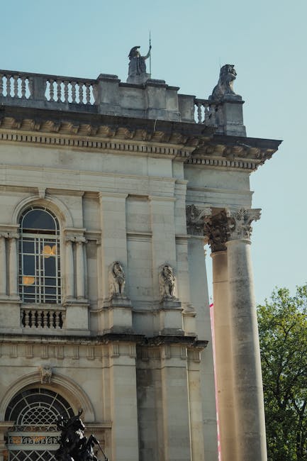 A close-up view of a historic stone building featuring classical architectural elements, including ornate decorative sculptures positioned on the roofline and a prominent fluted column supporting an entablature. The sculptures depict figures holding objects, and the top of the building has a balustrade with additional decorative statues. The photograph is taken during daylight with a partly cloudy sky and some greenery visible on the right side. This image highlights the architectural details typical of historic London structures, relevant to house removals and moving services such as those offered by Man and Van Pimlico. Such landmarks can often be part of relocation routes or serve as points of reference during home or office moves in the Pimlico area, supporting efficient planning of furniture transport and packing logistics.