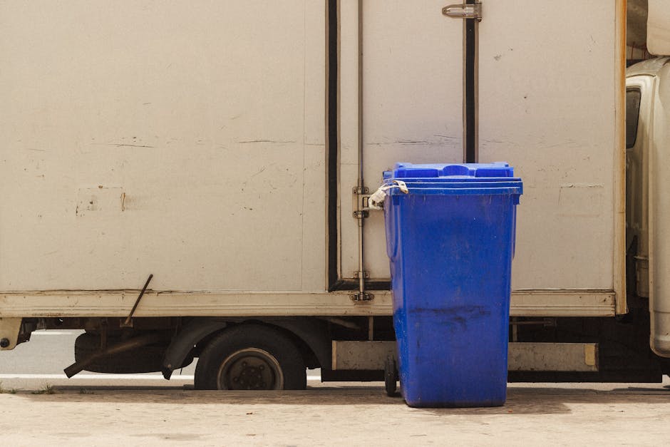 A small blue refuse collection truck with its bed tilted upward is in the process of unloading waste onto a large pile of mixed garbage, including plastic, paper, and organic debris, at an outdoor site surrounded by high, rocky, and uneven natural walls. Several people dressed in casual clothing, some carrying large white plastic bags, are manually collecting and sorting waste amidst the scattered trash. The environment appears to be a designated waste disposal area, with the truck positioned on a dirt ground and waste spilling from the truck's bed onto the heap. The scene reflects a waste unloading or disposal operation, which could be related to the waste management aspect of house removals or moving services, as handled by companies such as Man and Van Pimlico. Proper waste disposal procedures are crucial during home relocation and furniture transport to ensure compliance with legal requirements and environmental standards.