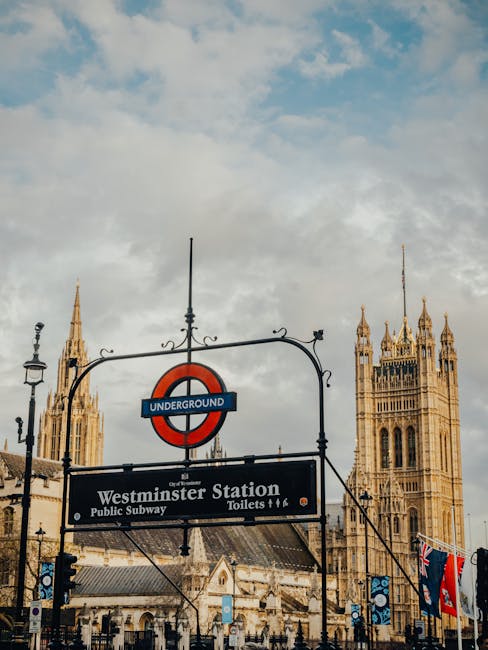 A street view showing Westminster Station with a sign for the London Underground overhead, positioned in front of the historic Palace of Westminster featuring ornate Gothic architecture and tall towers. The scene is outdoors during daytime with a partly cloudy sky, and the station sign indicates access to the public subway, toilets, and nearby amenities. This setting highlights an urban environment in central London, relevant for house removals and moving logistics. The presence of the station sign and historic building emphasizes the importance of location when planning home relocation or furniture transport services, as provided by companies like Man and Van Pimlico. The detailed architectural features and signage contribute to a comprehensive visual understanding of the area involved in moving and packing processes within the cityscape.