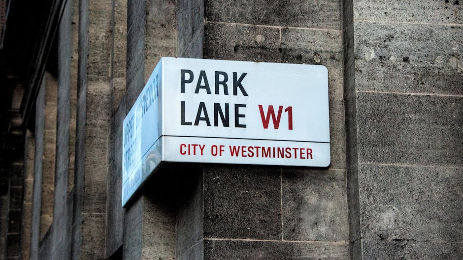 A close-up photograph of a white rectangular street sign mounted on a dark stone wall in Westminster, London. The sign displays the words 'PARK LANE W1' in bold black and red letters, with 'CITY OF WESTMINSTER' printed in red beneath. The sign is attached to a wall with visible textured stonework, and on the left side, a part of a metal railing or fence can be seen. This street sign indicates a location within a central London area commonly associated with upscale addresses and is relevant in the context of house removals and moving logistics, especially when obtaining permits for parking or loading during home relocation procedures. The lighting appears natural, emphasizing the clarity and prominence of the sign against the textured background.